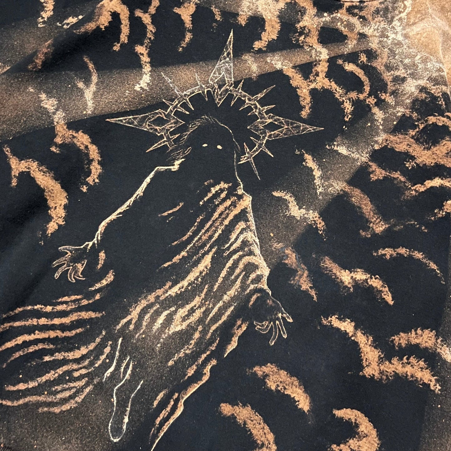 A close-up of a black crewneck sweatshirt featuring a distressed, bleach-splattered graphic of a hooded figure with a star-like crown, surrounded by abstract splattered patterns in a warm tan/beige color.